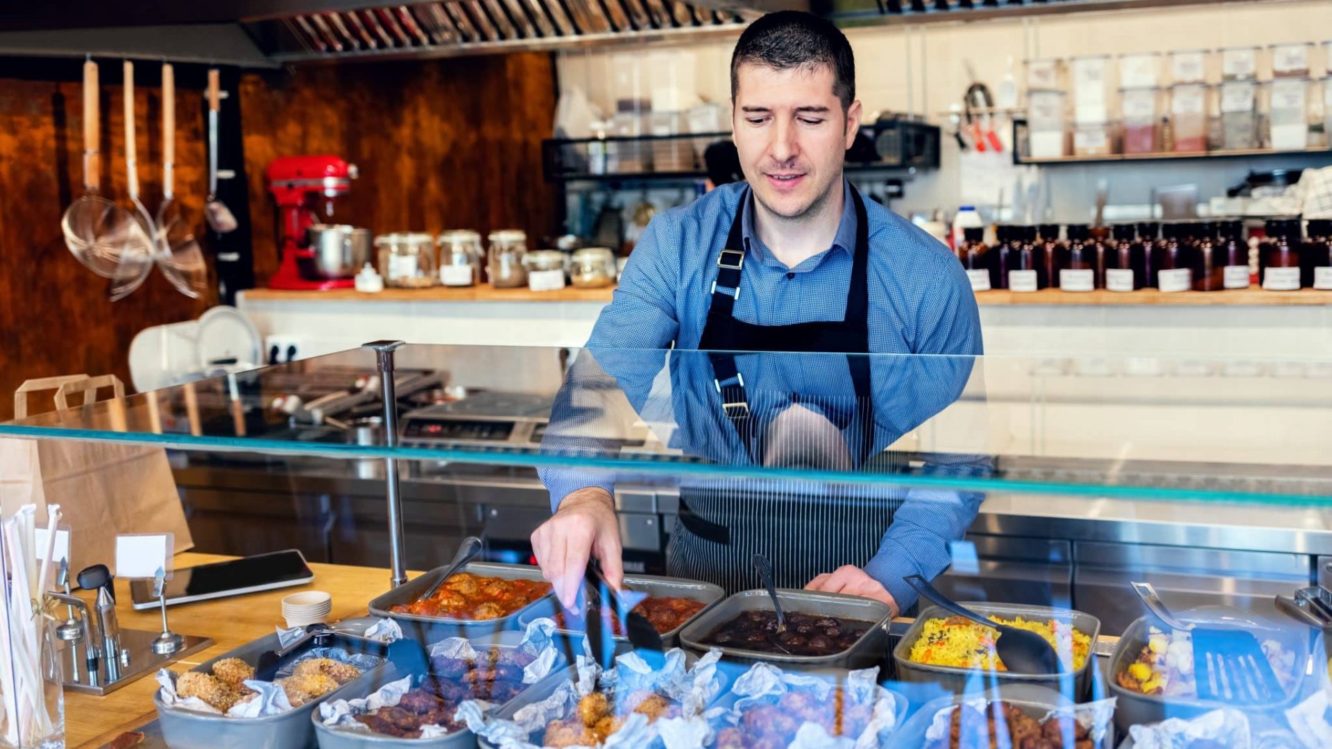 Happy waiter wearing apron serving takeaway food to customer at counter in small family restaurant.