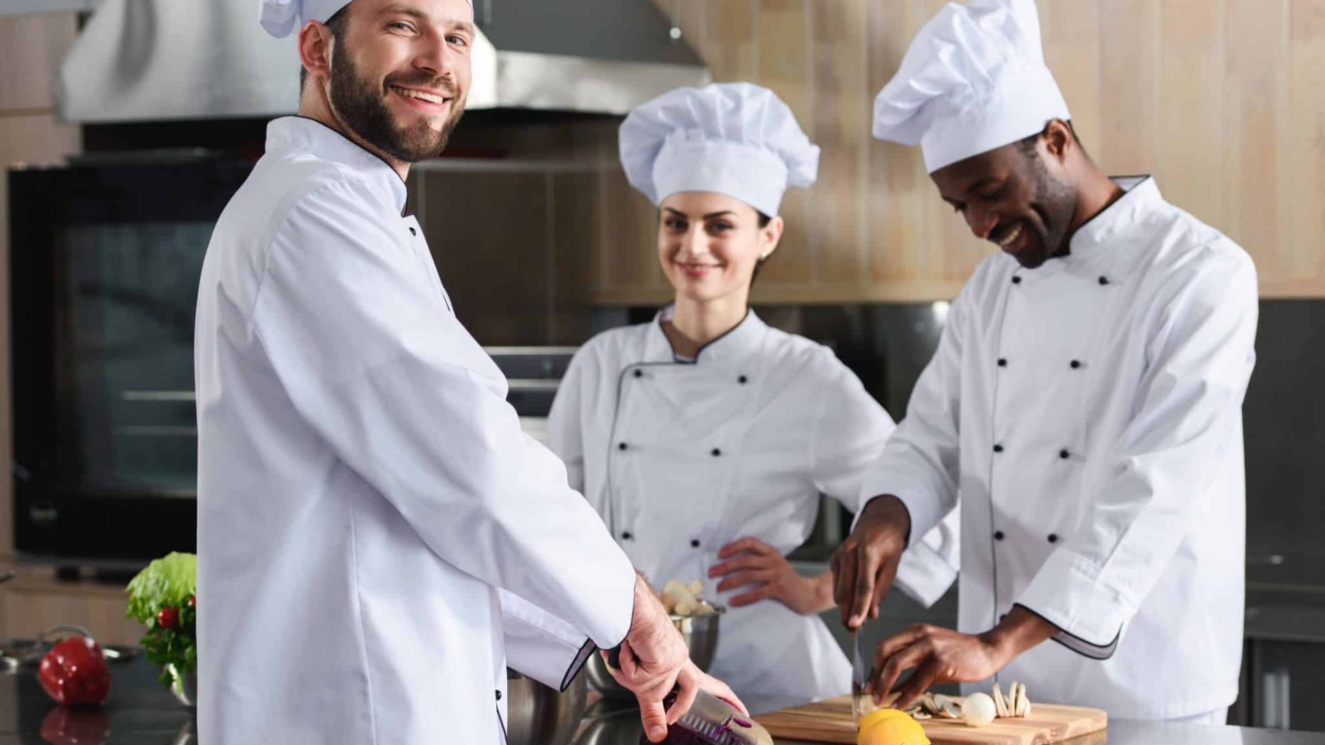 Diverse team of cooks smiling while cutting ingredients by kitchen table